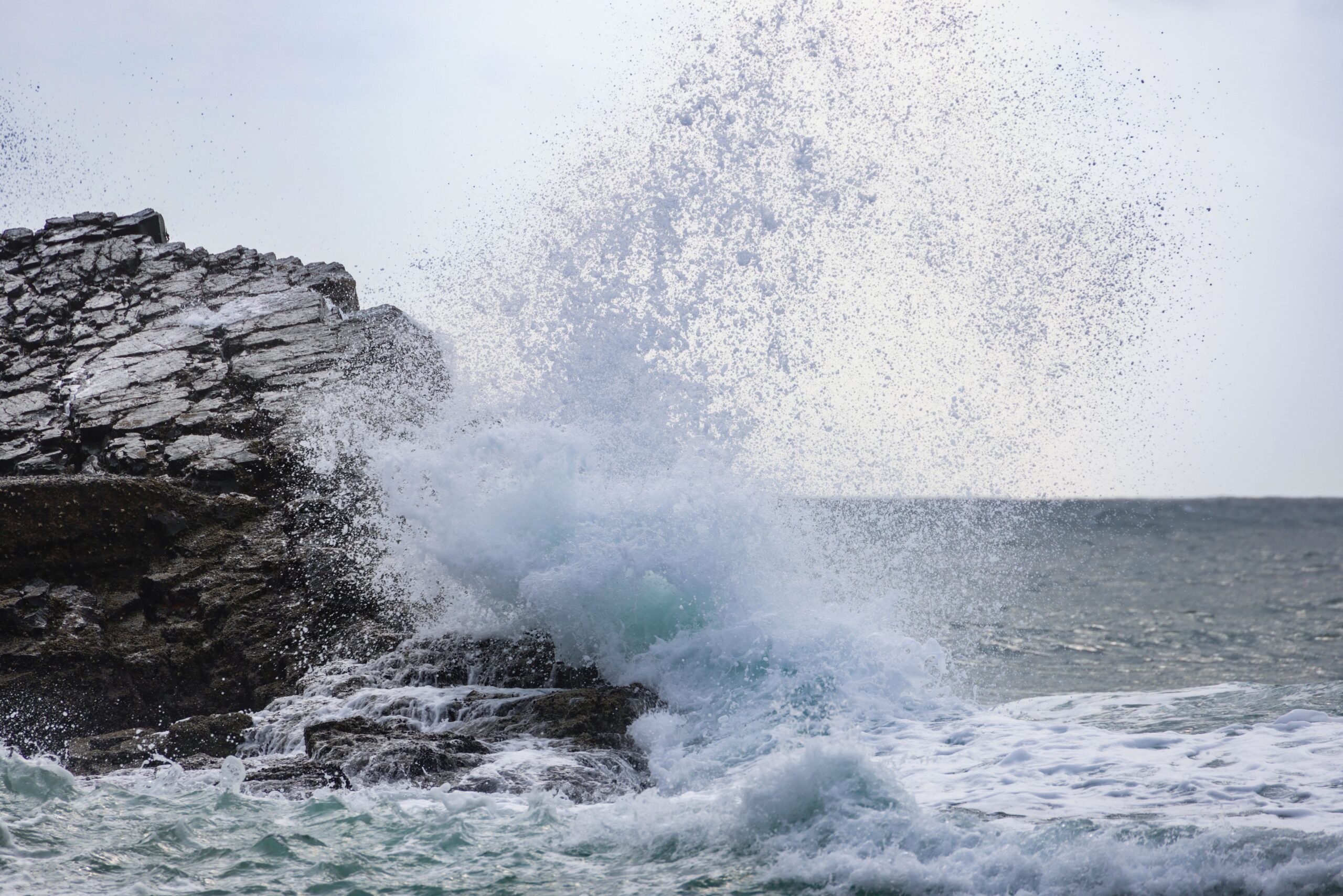 A powerful ocean wave crashes against dark coastal rocks, sending white spray high into the air.